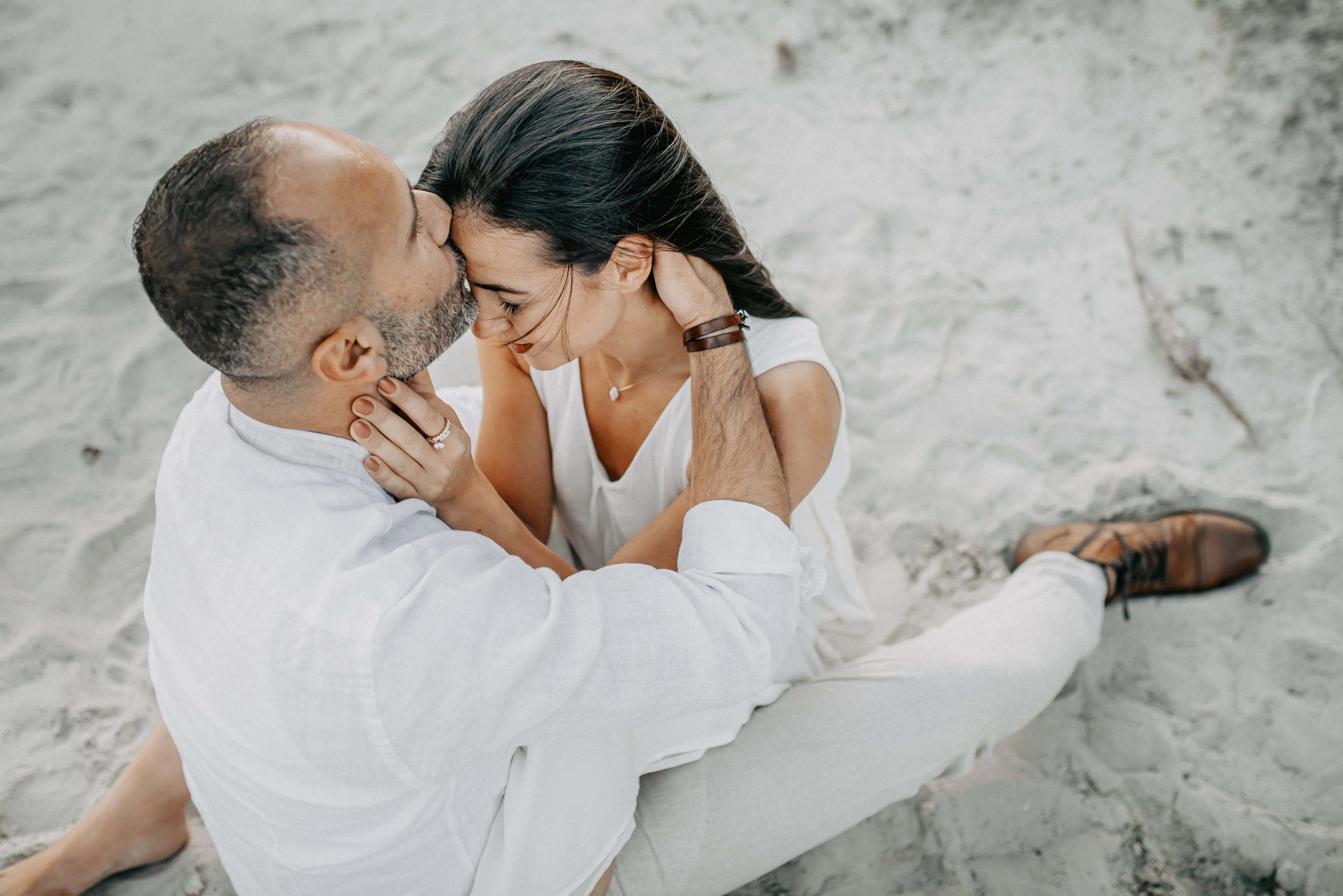Im Sand der Heide-Landschaft eng umschlungen sitzend, küsst ein Mann seiner Frau auf die Stirn, während er ihren Kopf mit beiden Händen und sie ihre Augen geschlossen hält.