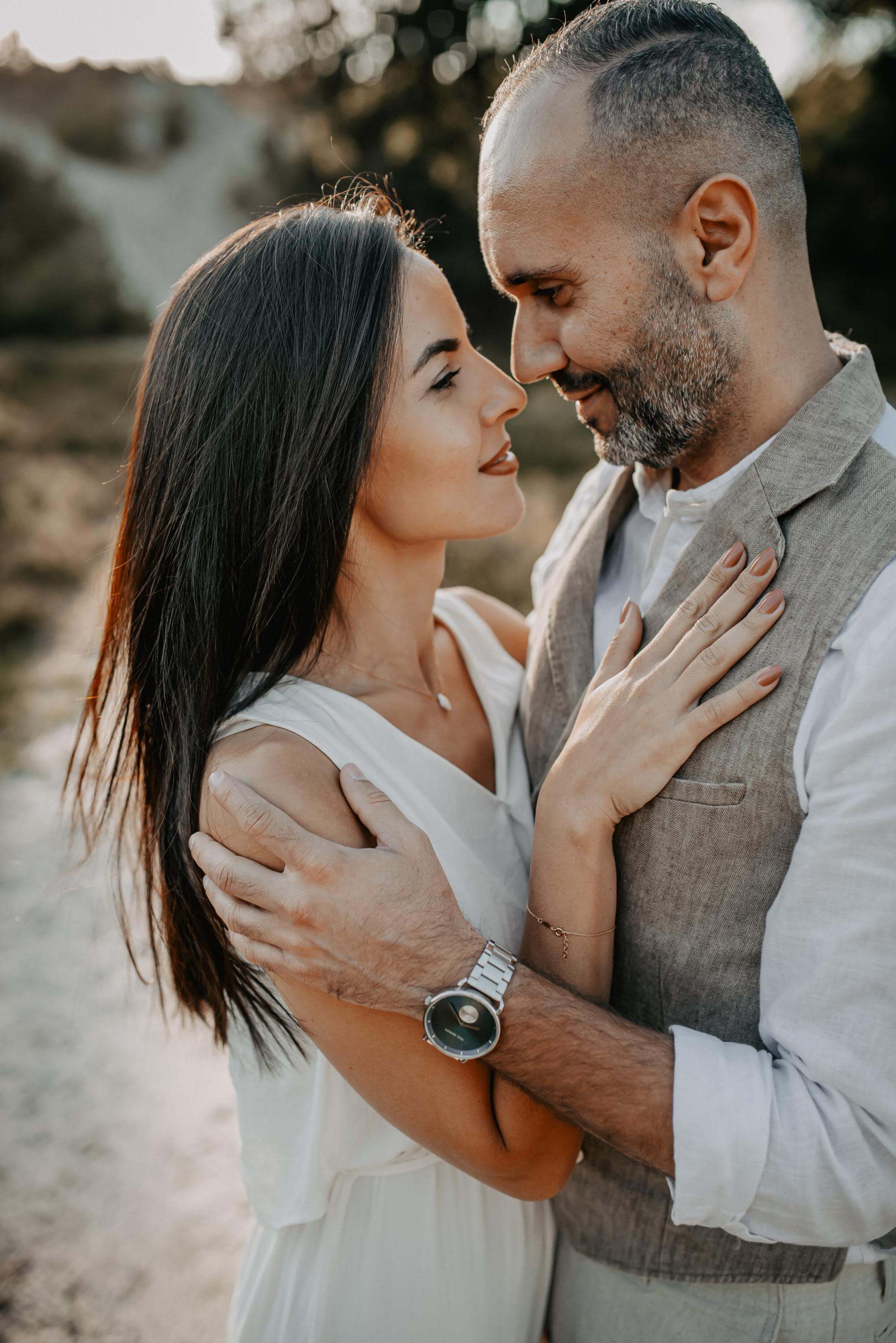 Standing nose-to-nose close together, a woman and man look into each other's eyes in love for an engagement photo. Both are dressed in southern vintage style, with him wearing a beige linen waistcoat.