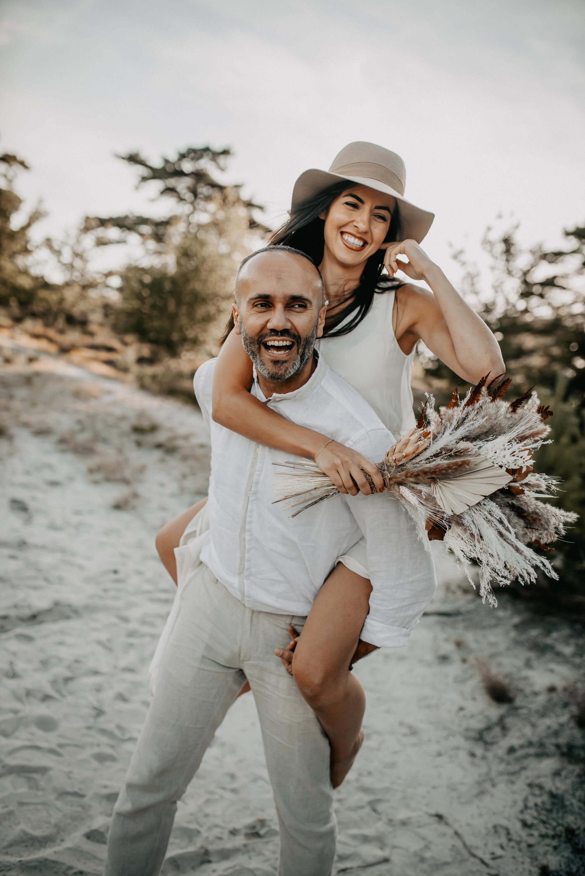 In a relaxed mood, a man carries his wife in a cowboy hat piggyback on his back while they both laugh in the sandy heathland.