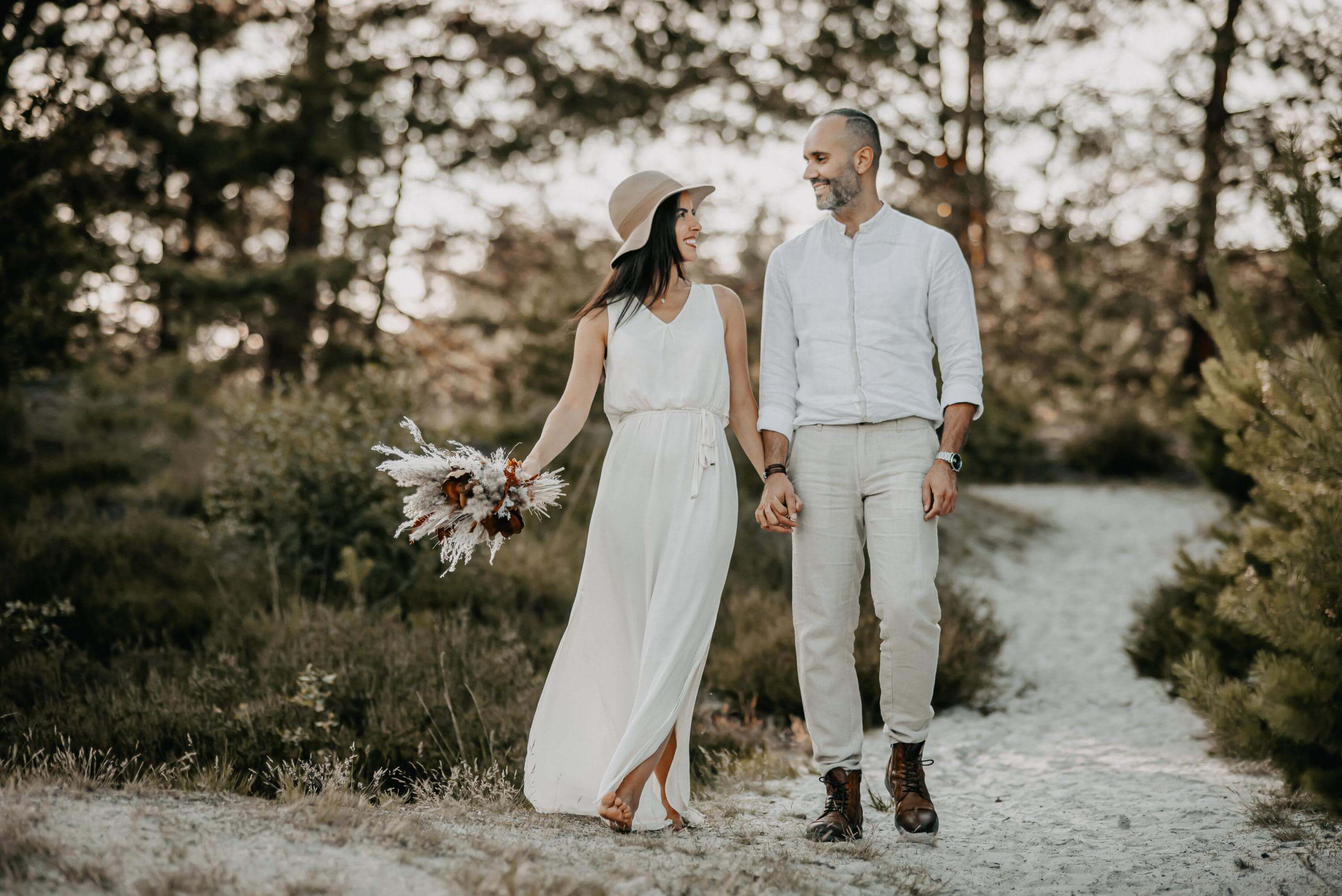 For an engagement photo shoot, a couple of lovers walk holding hands and looking at each other in love through a sandy landscape of paths with a forest. She is still holding a bouquet of dried flowers
