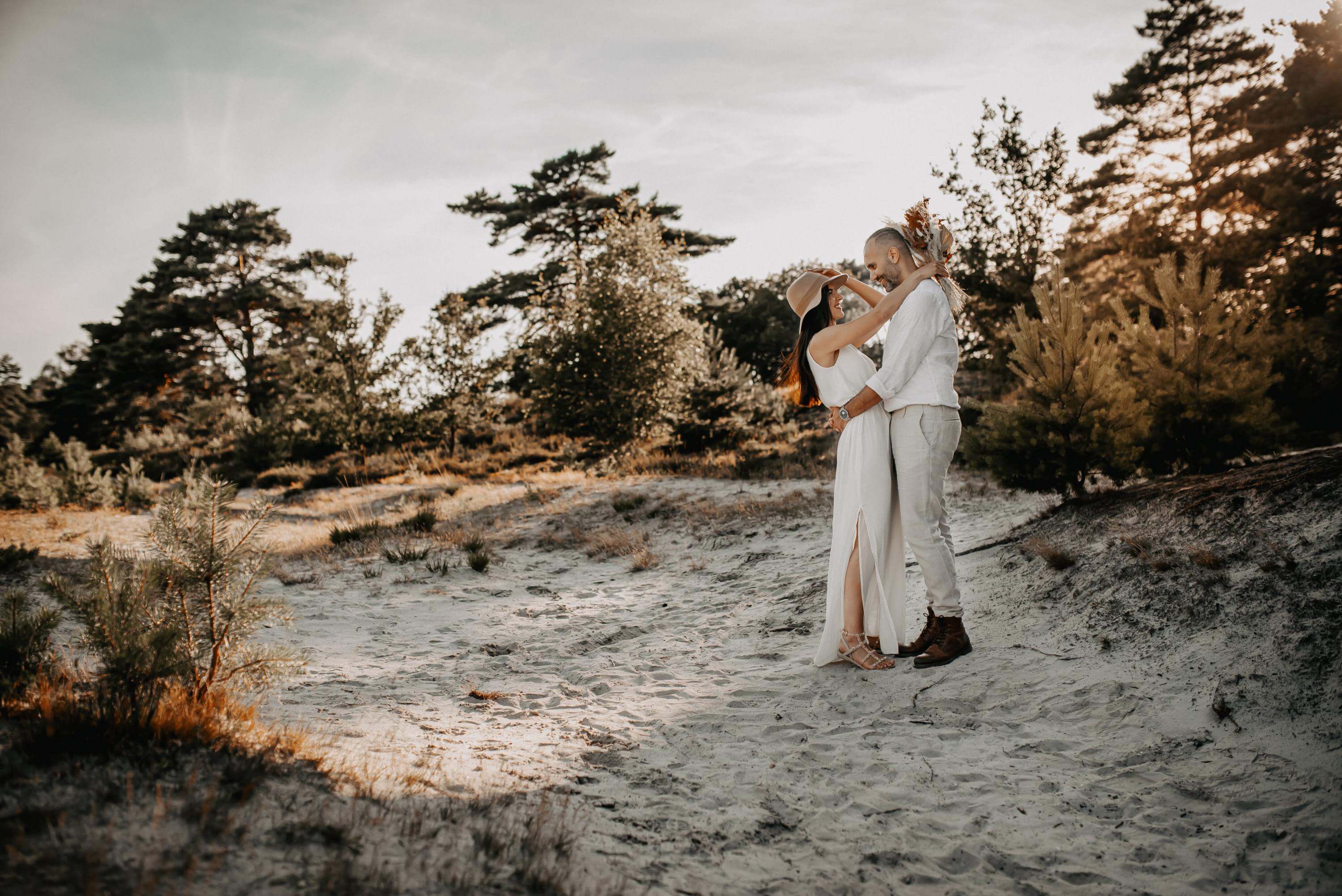 A pair of lovers, wearing light beige clothing, stand on a sandy dune in a heath landscape in the flat sun. They both hug and look into each other's eyes.