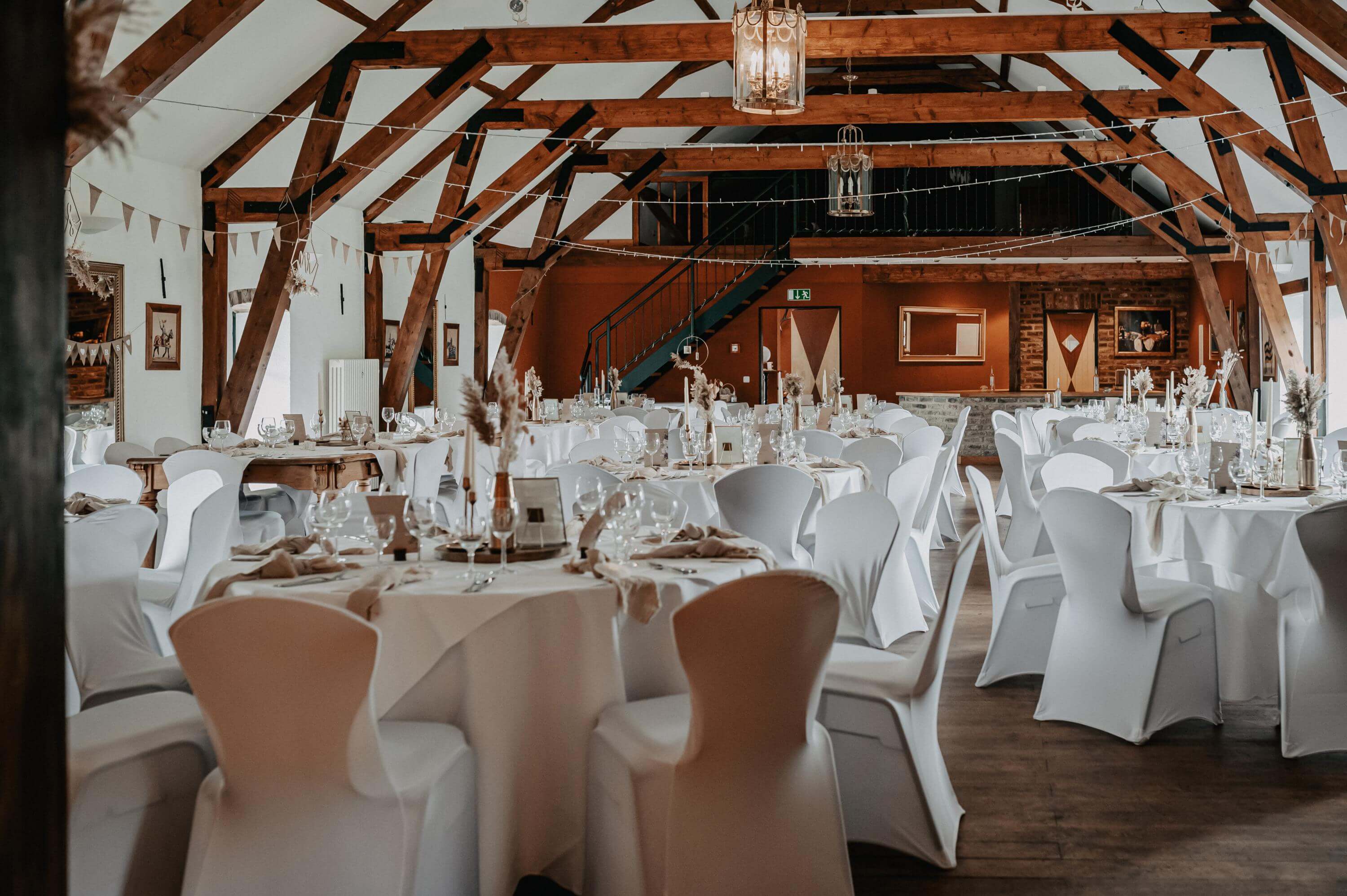 Die große helle Scheune mit Holzbalken der Hochzeitslocation La Maison Blanche  ist mit runden Esstischen und Stühlen mit weißen Hussen im Vintage Stil ausgeschmückt.