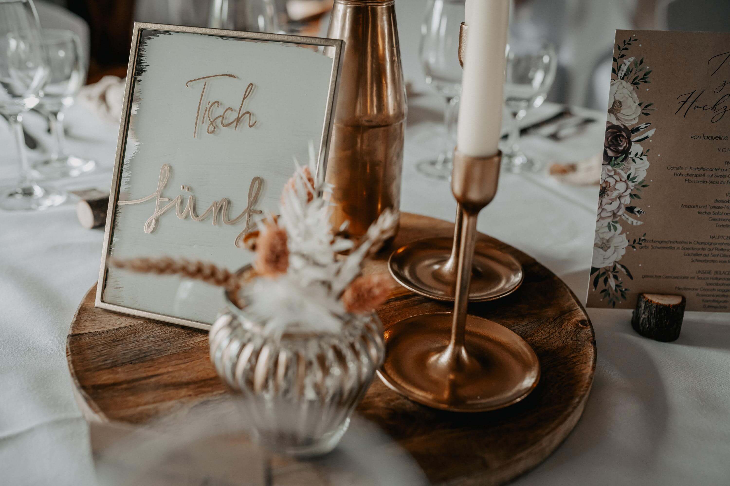 Die kupfer- und weißfarbene Tischdekoration mit Tischnummer im Vintage Stil wirkt edel und modern mit den eisernen Kerzenständern und Trockenblumen auf einem Holzteller kombiniert.