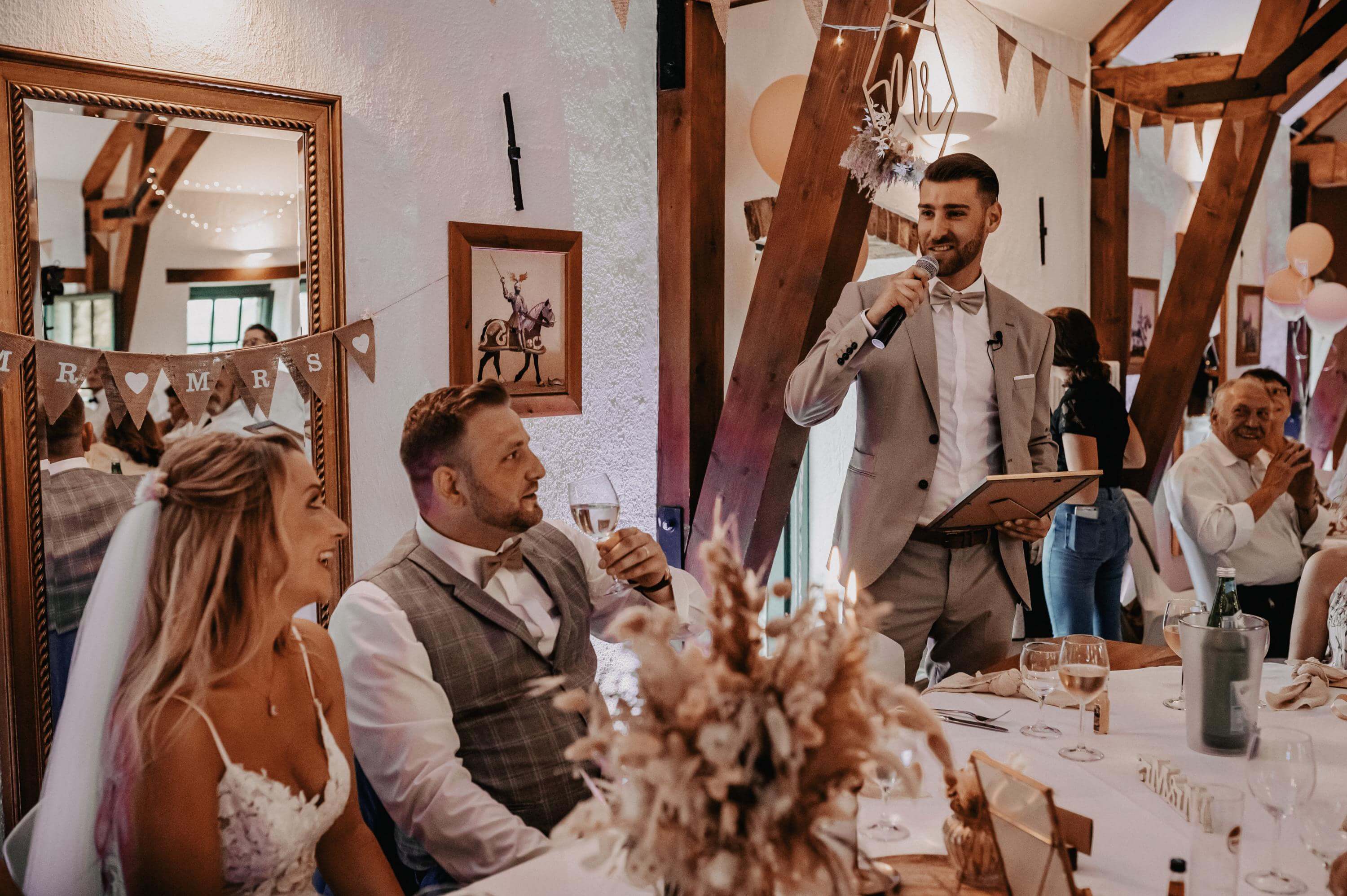 At a wedding reception, a best man stands at the table of the groom, who is seated next to him, holding a picture in the frame. He speaks a touching speech with the microphone that brings tears to the eyes of the bridal couple.