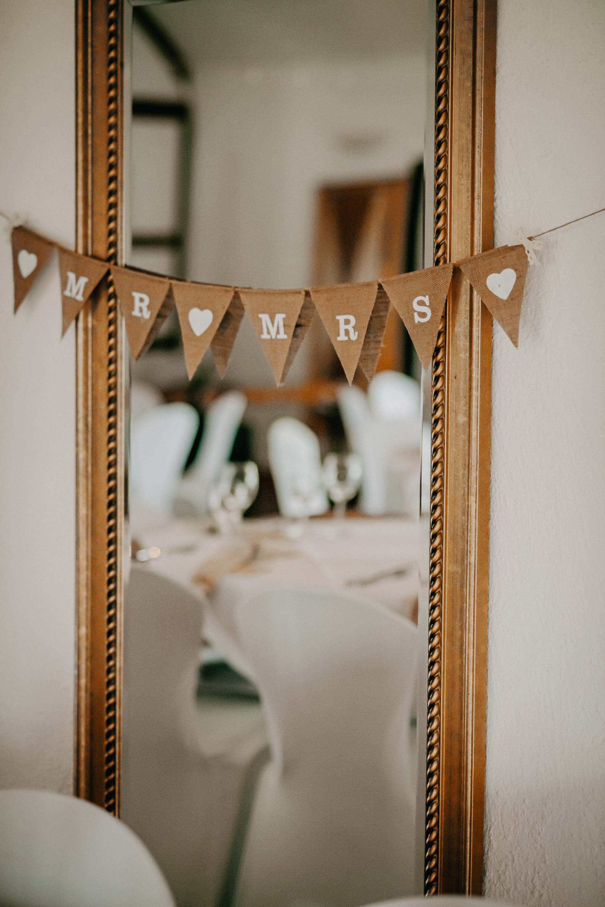 Vor einem großen langen Spiegel mit prunkvollem Rahmen an der Wand hängt eine Wimpelkette mit dem Motiv 'Mrs & Mr' zur Hochzeit. Im Spiegel sieht man einen Stuhl mit weißer Husse an einem runden Tisch für die Hochzeitsgesellschaft.