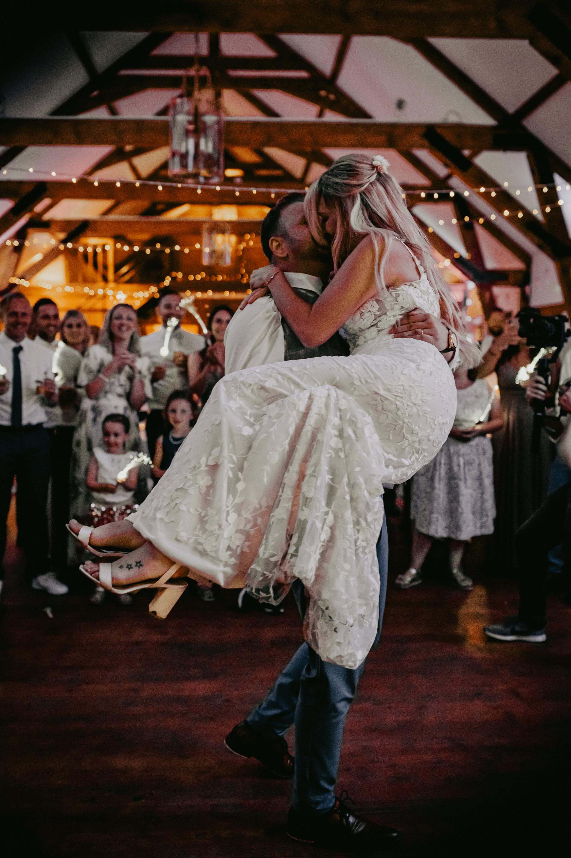 Beim Paartanz in der romantisch geschmückten Scheune mit Lichterketten tanzt das Hochzeitspaar inmitten der Hochzeitsgesellschaft. Er trägt sie auf seinen Armen und beide küssen sich freudig.