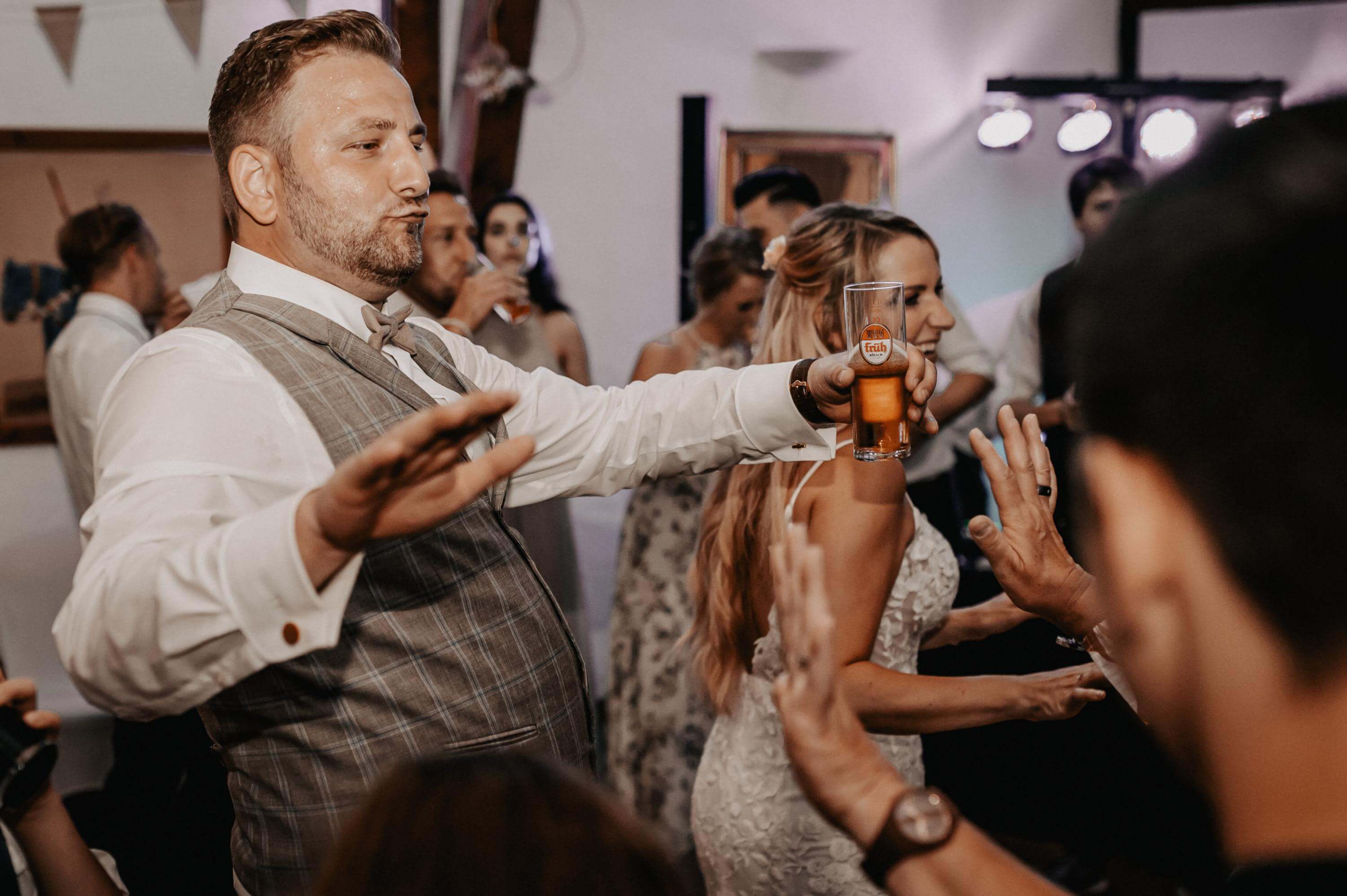 Ausgelassen mit Bierglas in der Hand tanzt der Bräutigam neben der Braut am späten Abend auf der Tanzfläche in einer Scheune zusammen mit der Hochzeitsgesellschaft.