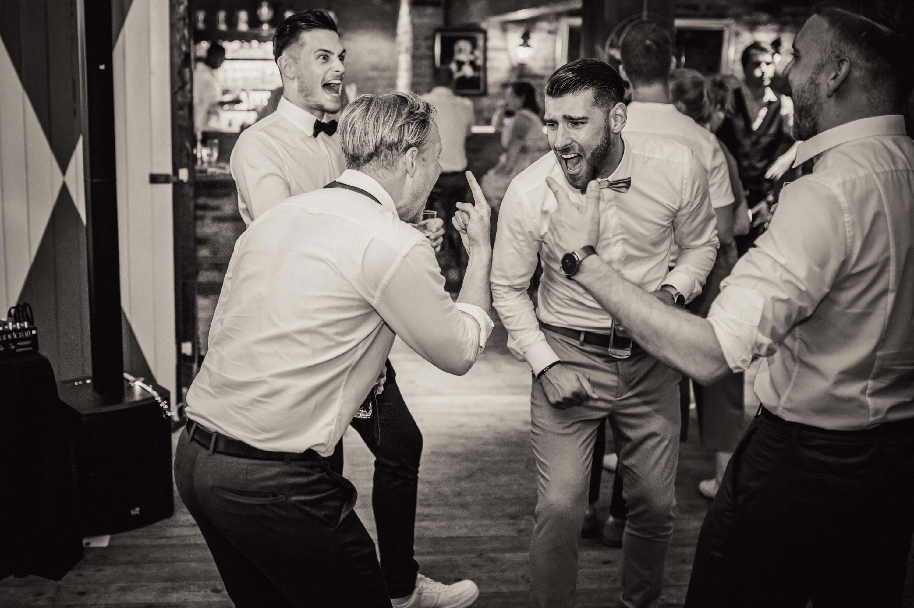 Extrem gelöst und aufgedreht tanzen junge Männer in hellen Hemden und mit freudig aggressivem Gesichtsausdruck bei einer Hochzeit frei nebeneinander.