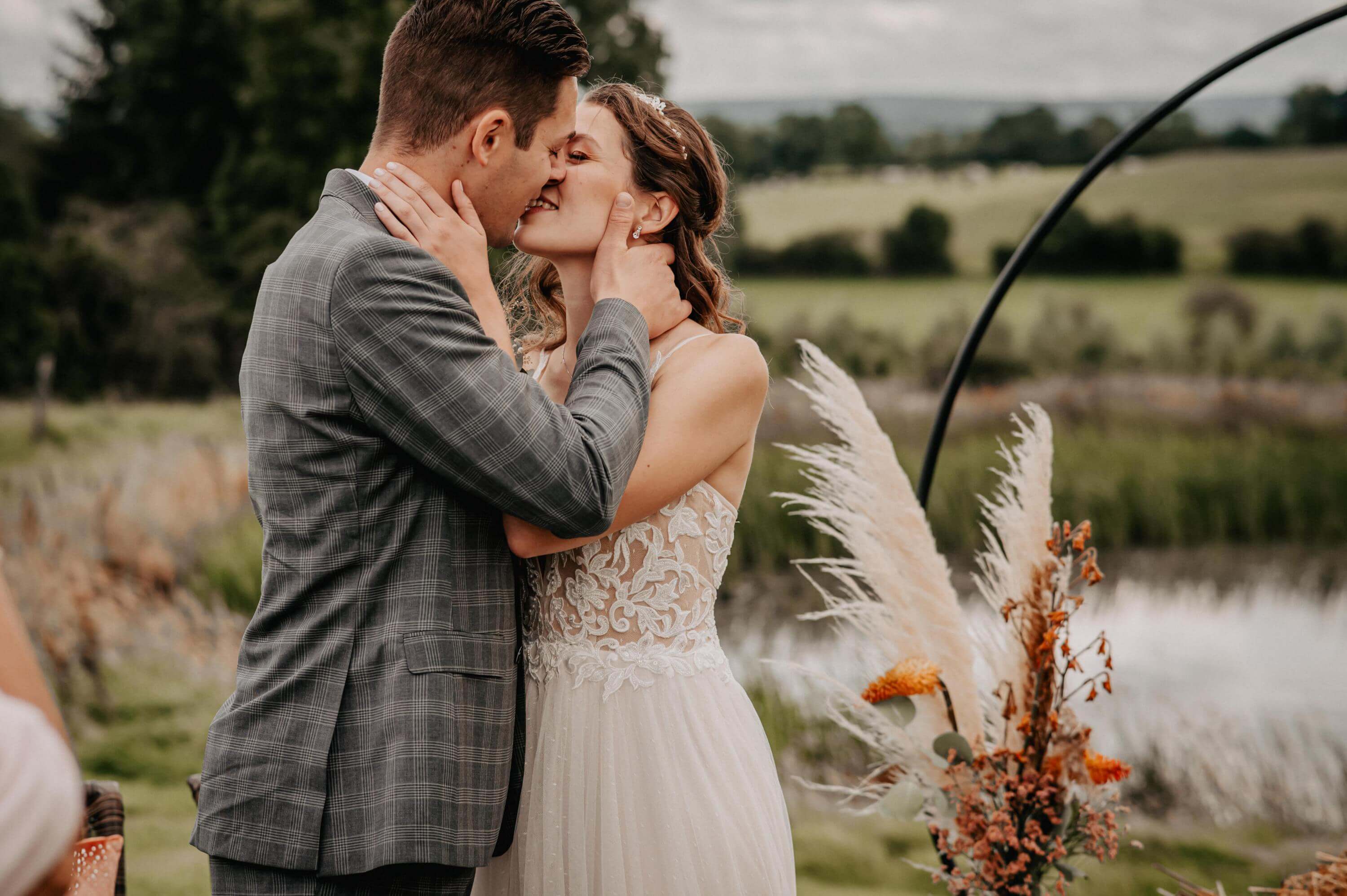 The close-up shows a wedding couple gently kissing with closed eyes at a boho-style outdoor wedding in front of a small lake and a large, round wedding arch.
