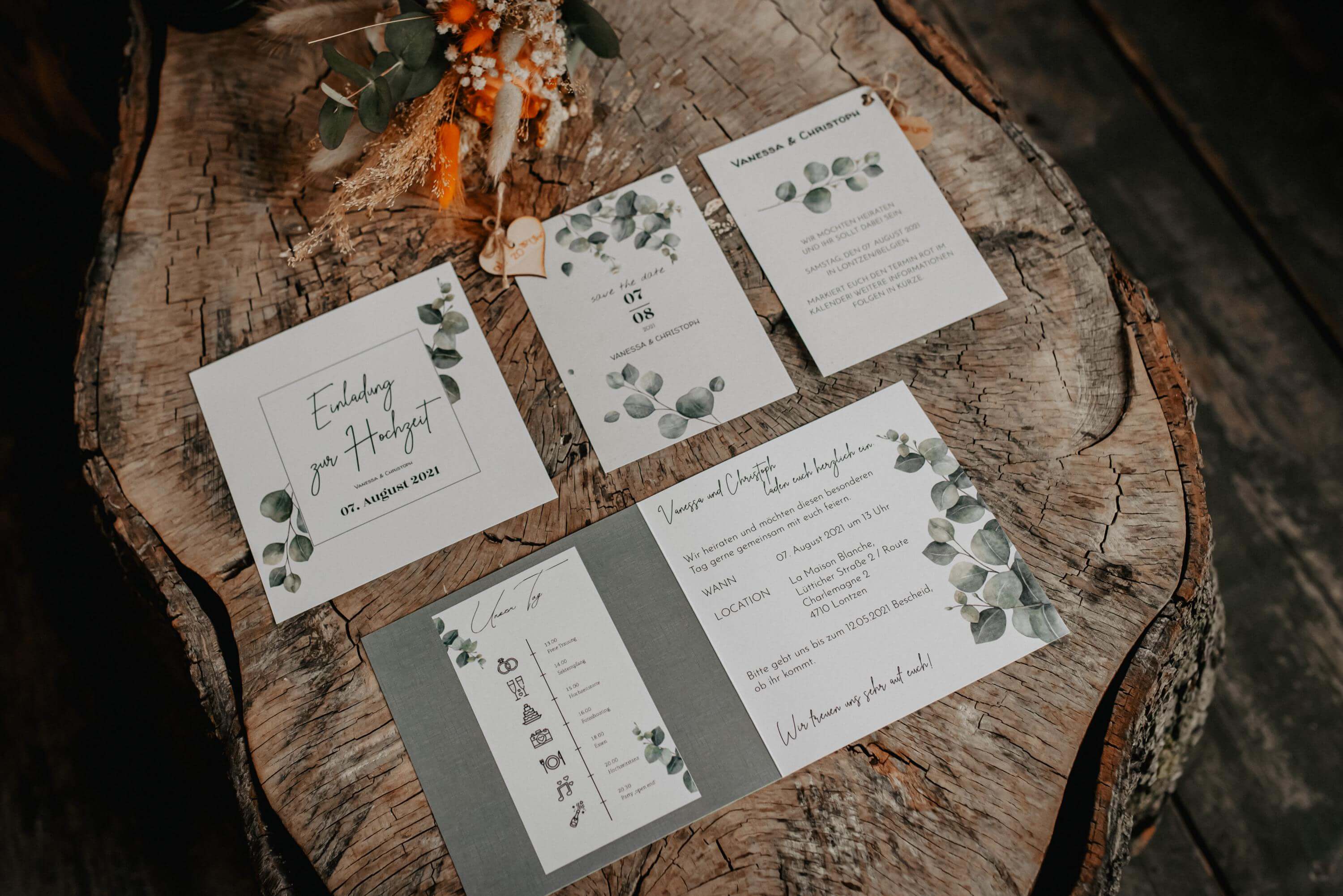 All the stationery for a wedding in a eucalyptus-colored design with 'Save the Date', invitation, menu and schedule is spread out on a tree stump.