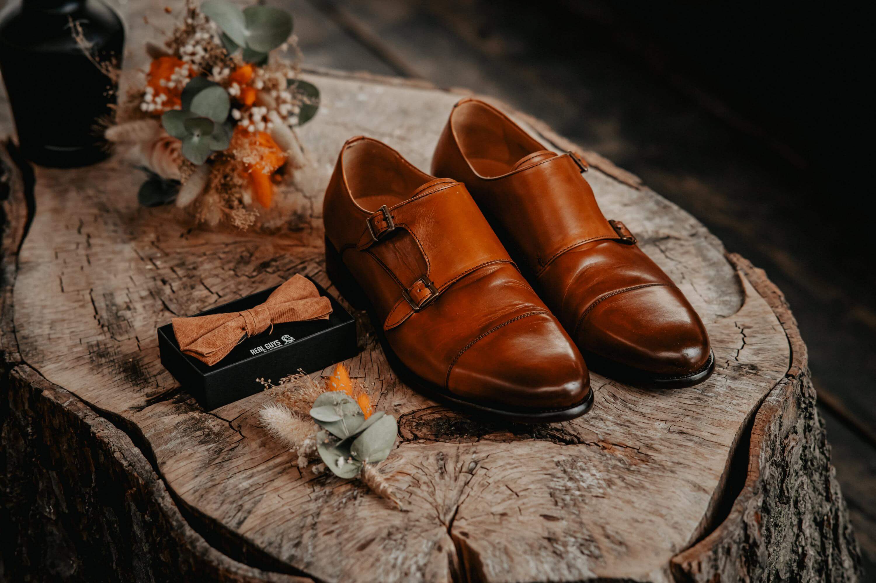 The groom's brown leather shoes and his noble brown fabric bow tie with brooches made of dried flowers are on a tree stump at the Getting Ready.