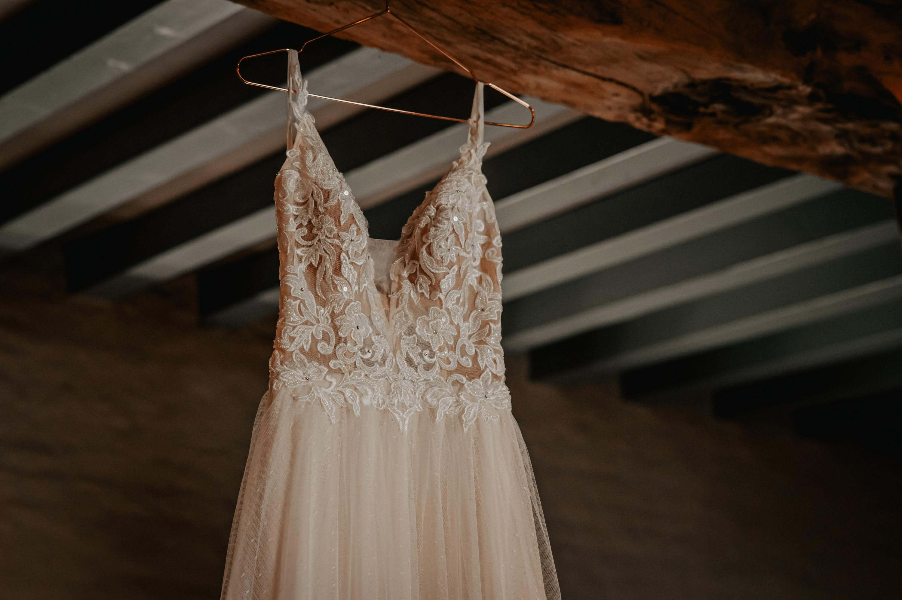 A light, slightly glittering wedding dress with coarse floral lace and narrow straps hangs on a copper-colored hanger from the ceiling beam of a hotel room.