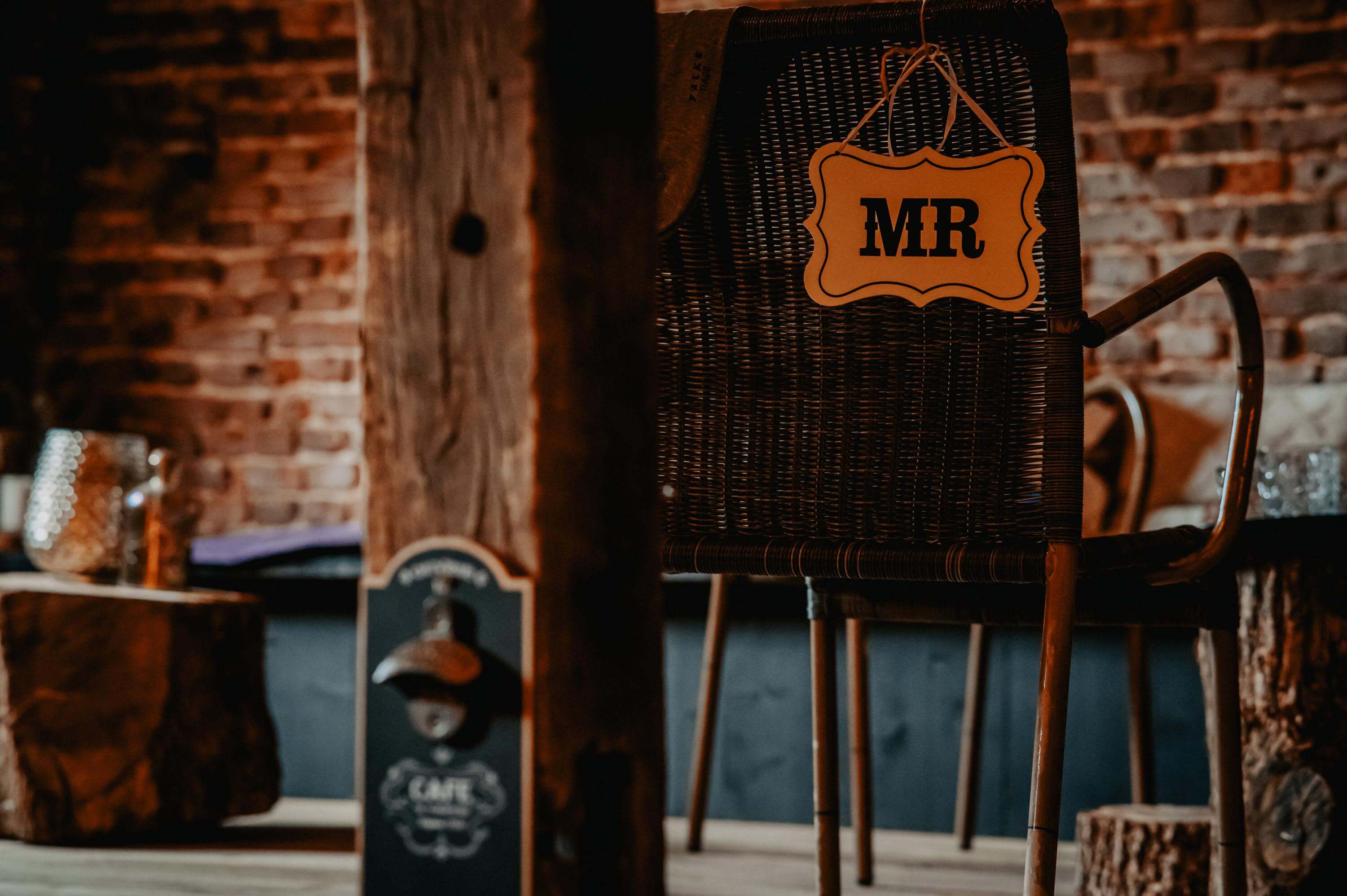 Hinter einem dunklen Stützbalken aus Holz steht der geflochtene Korbstuhl mit einem Anhängeschild 'Mr' bereit für das Getting Ready des Bräutigams.
