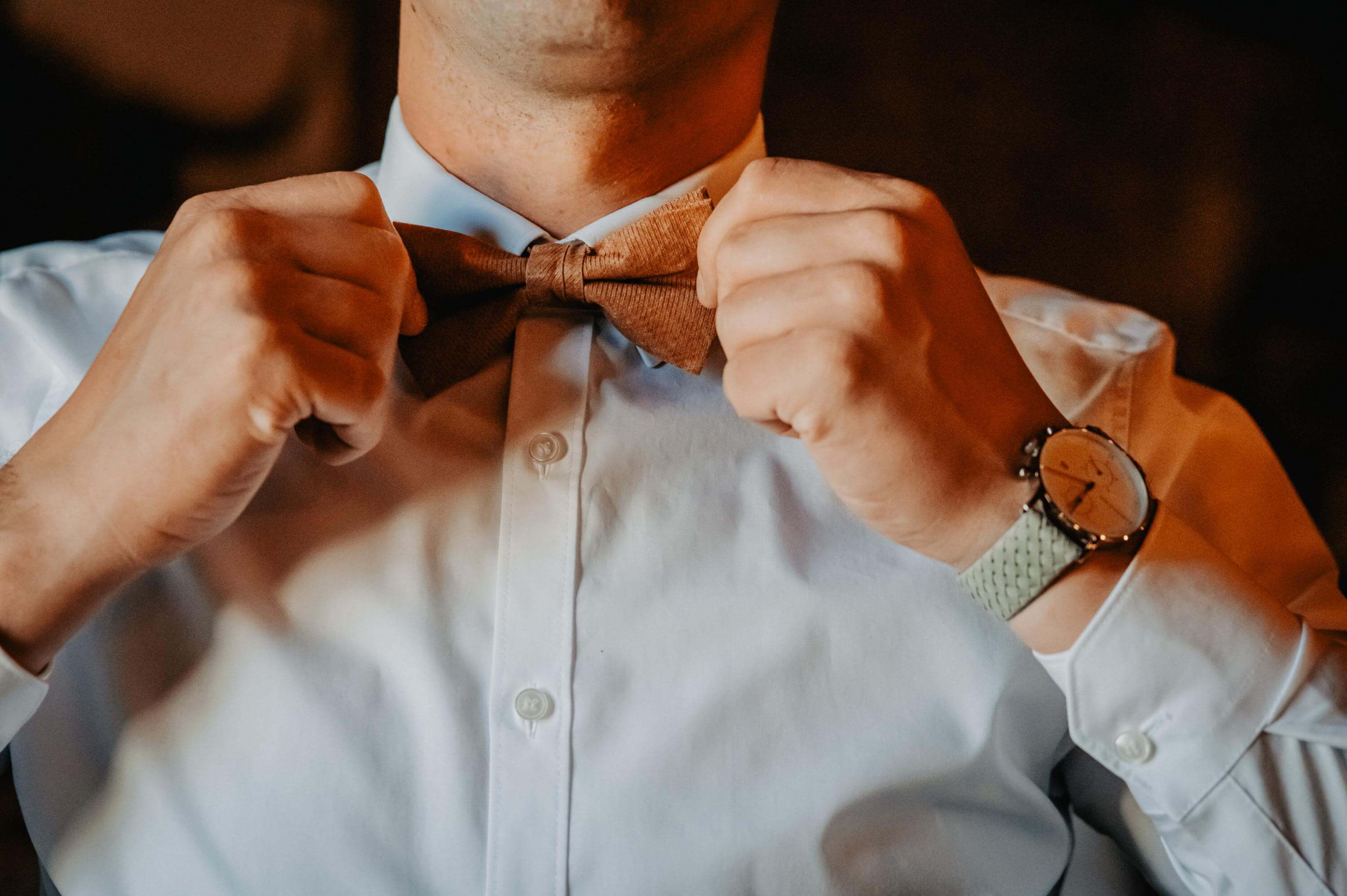 Klassische Nahaufnahme beim Getting Ready des Oberkörpers des Bräutigams im weißen Hemd und mit Armbanduhr, der sich seine Fliege am Hals mit beiden Händen gerade richtet.