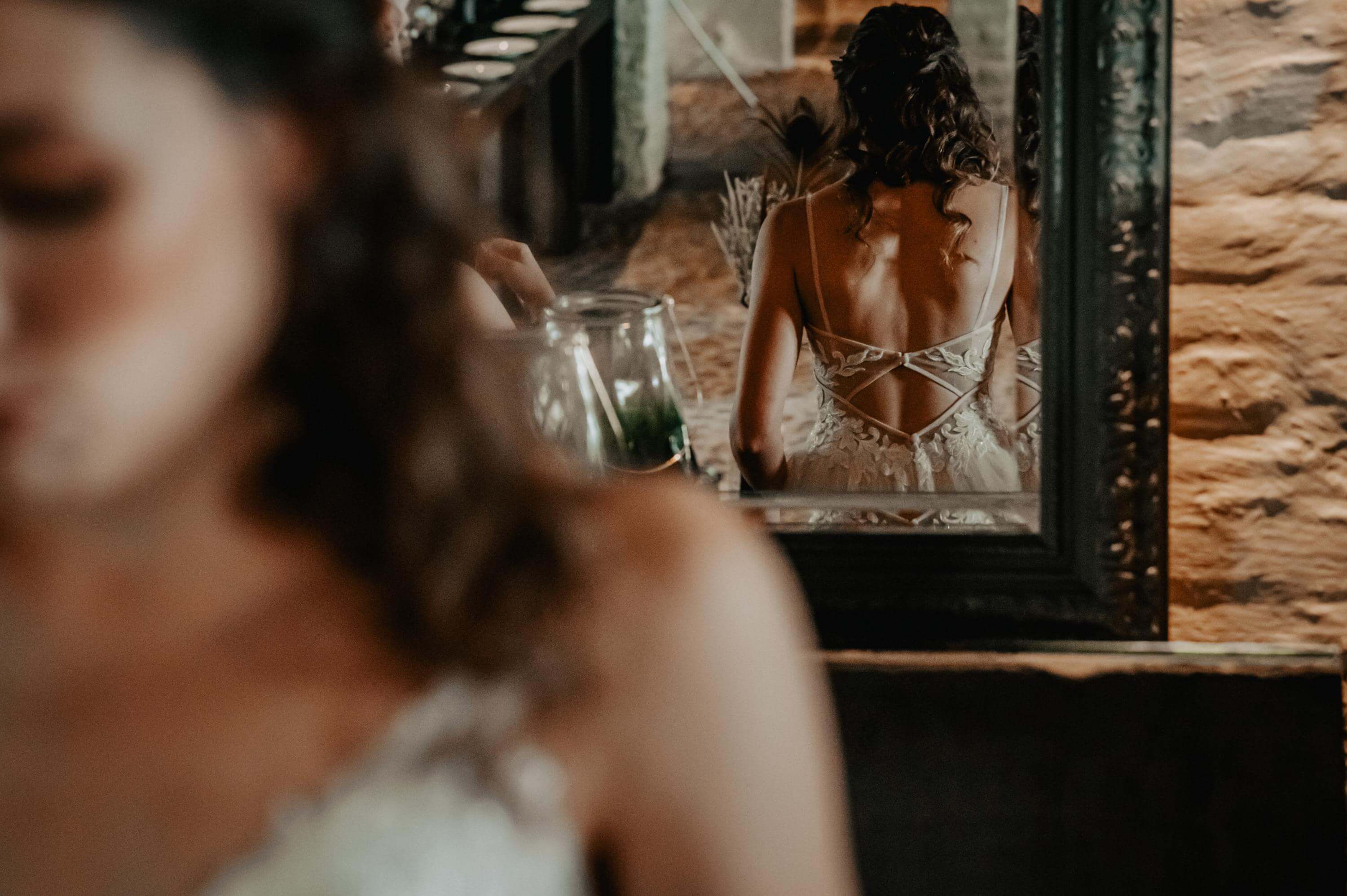 In a large mirror you can see the rear view of the bride in a wedding dress with curly hair and a wide back décolleté.