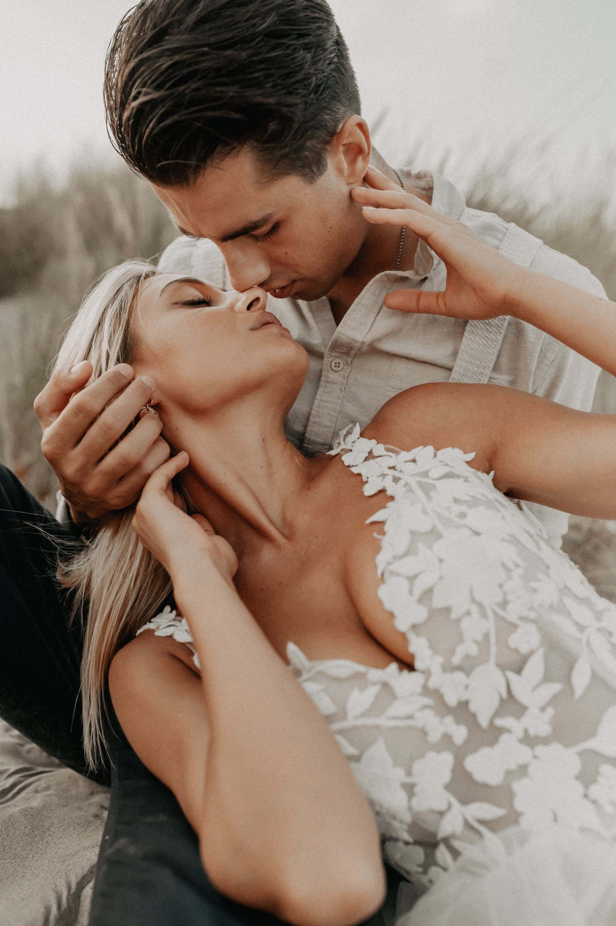 A woman in a wedding dress lies on the lap of her man in a vintage linen shirt during an after-wedding shoot on the beach. Both look deep into each other's eyes while showing off her deep, sexy cleavage.