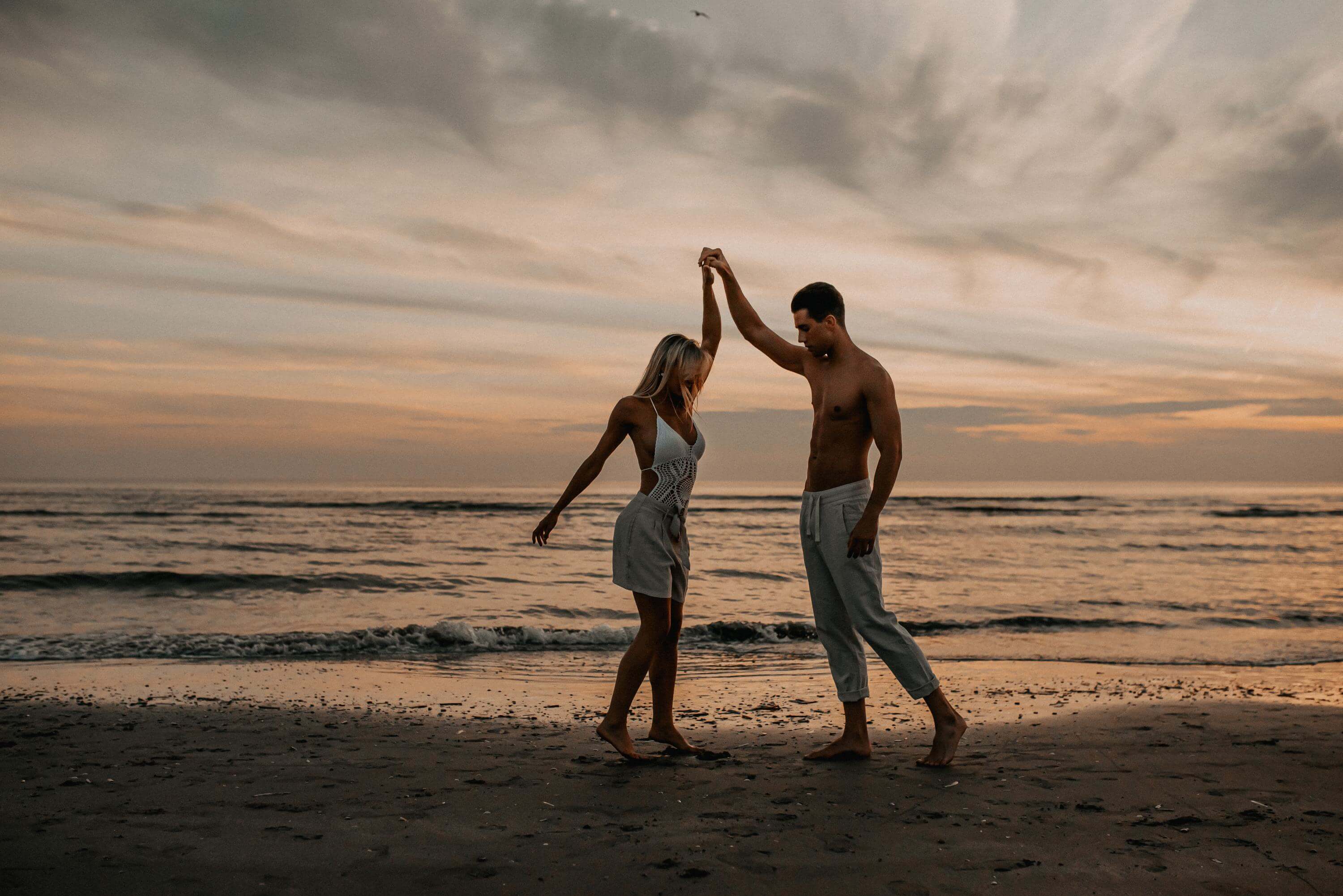 Bei Sonnenuntergang am Meer tanzt ein junges Paar mit sommerlich sexy Vintage-Outfit am Strand.