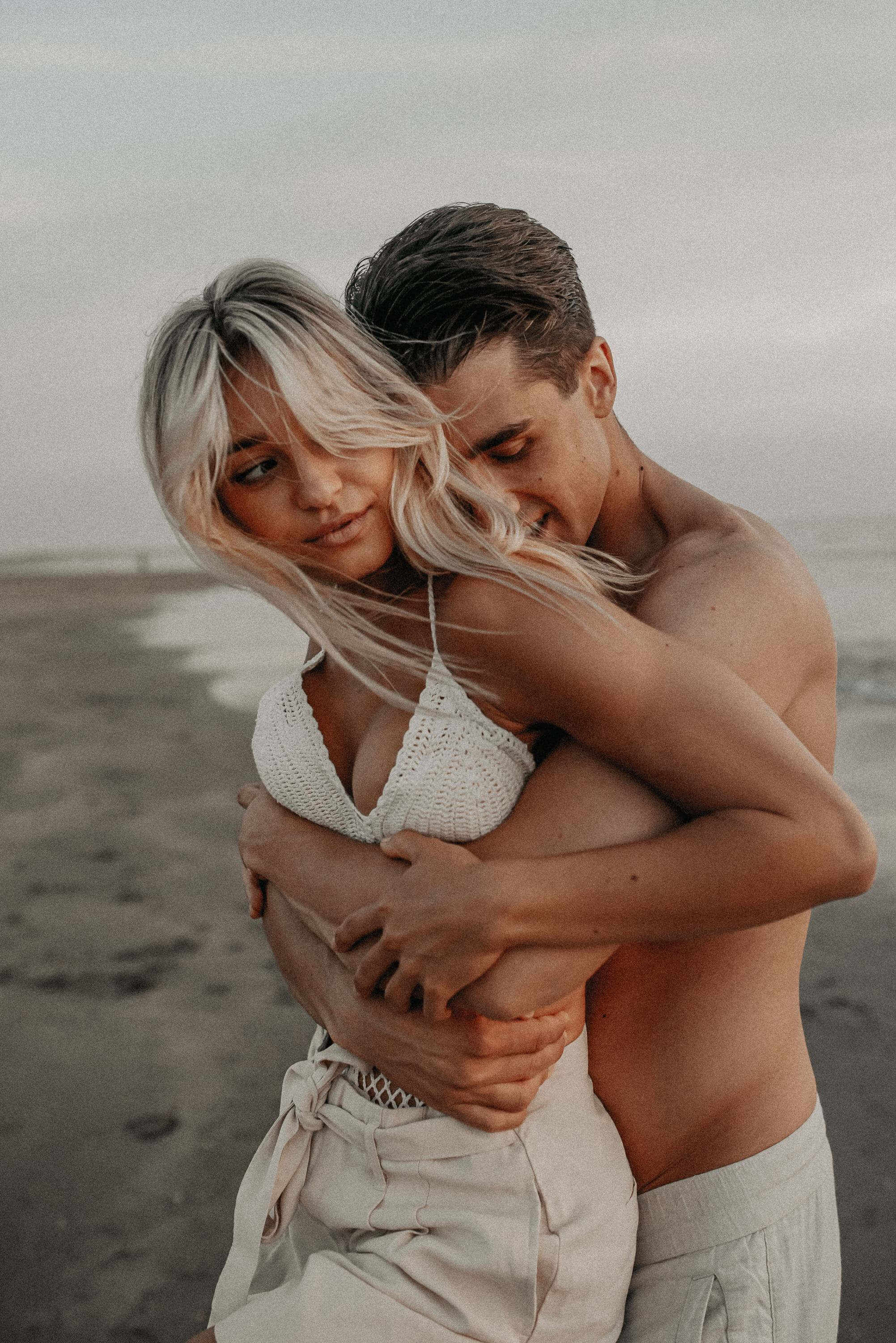 Seine Arme von hinten um sie geschlungen hält ein junger Mann mit freiem Oberkörper seine mit einem Vintage-Oberteil gekleidete Frau am Strand liebevoll fest.