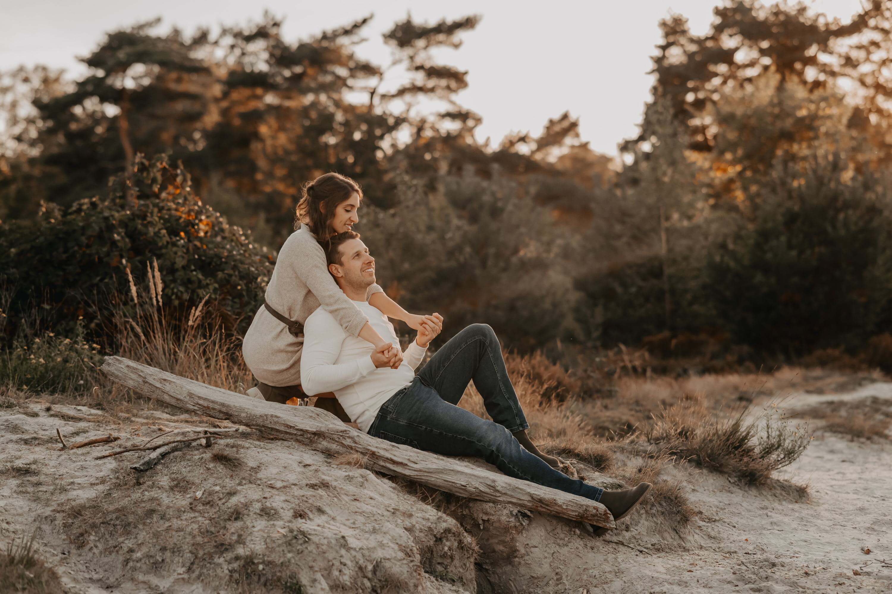 Hinter einem Baumstamm im Heidesand sitzend verbringt ein Liebespaar eine romantische Zeit Händchen haltend am See.