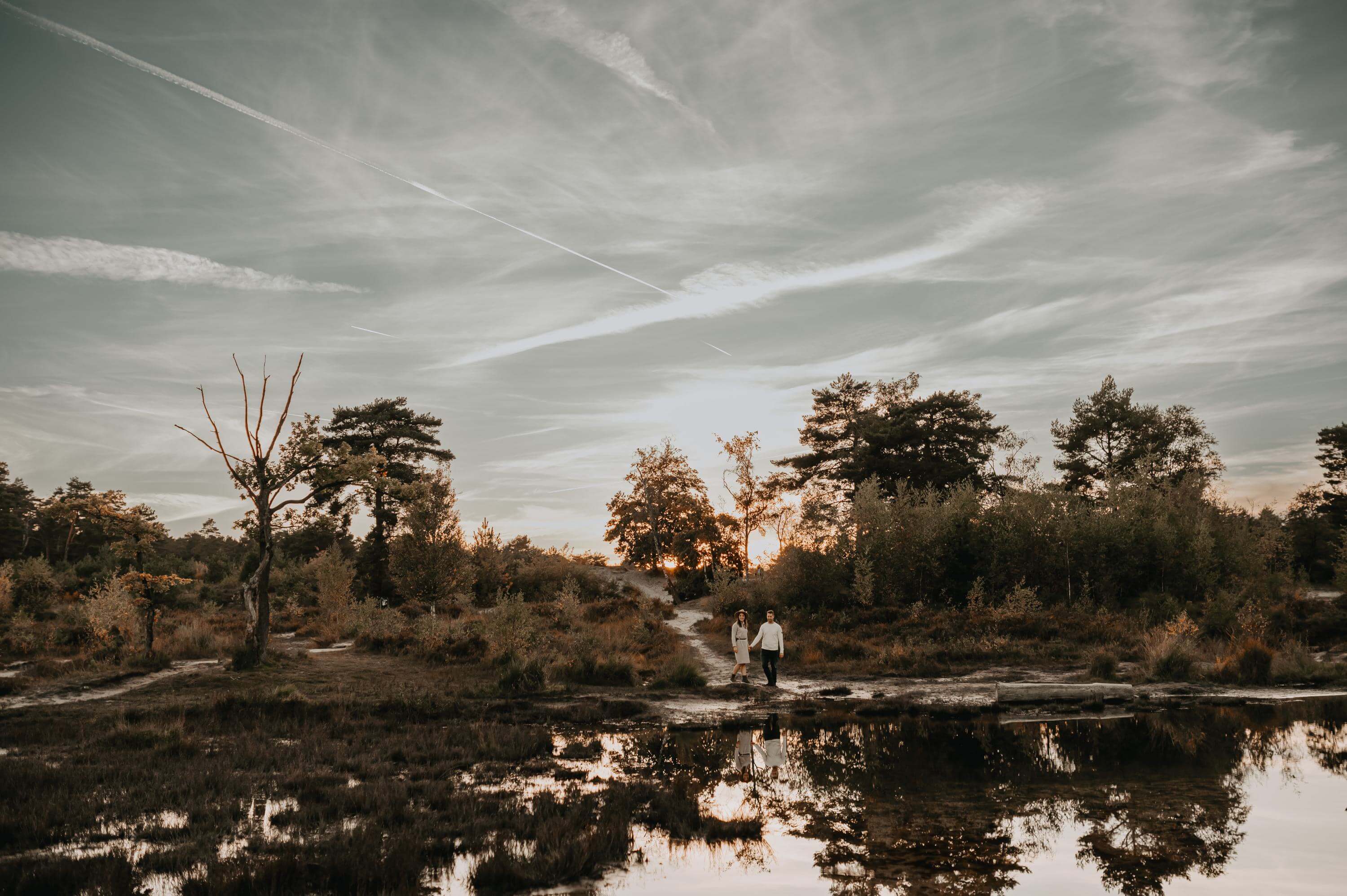 Im Sonnenuntergang steht ein Liebespaar Hand in Hand an einem See in der Heide. Beide spiegeln sich in der Wasseroberfläche.