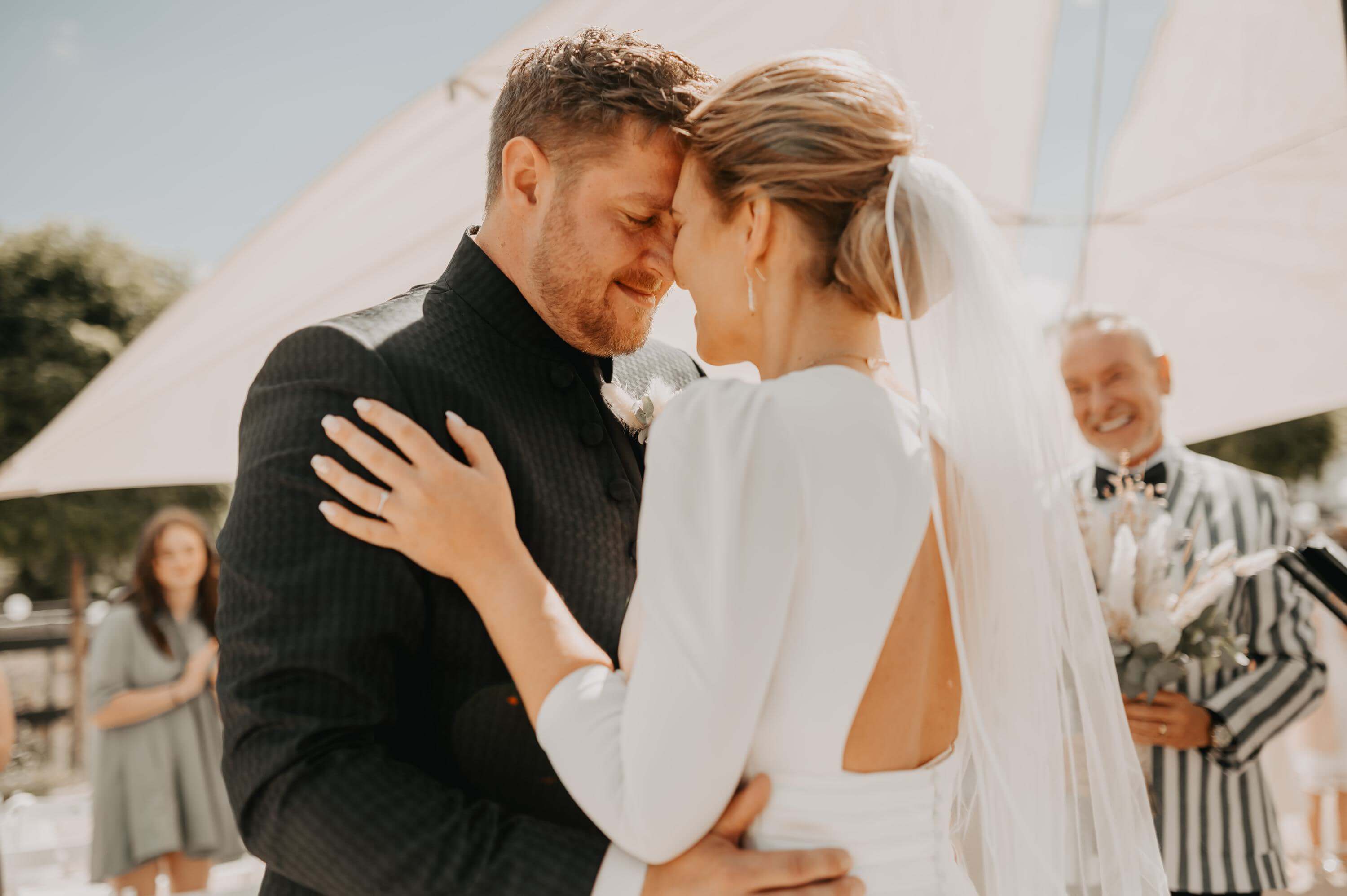 During the speech of the wedding speaker at a free wedding ceremony in the sun, the bridal couple face each other forehead to forehead. Both are smiling and have their eyes closed.