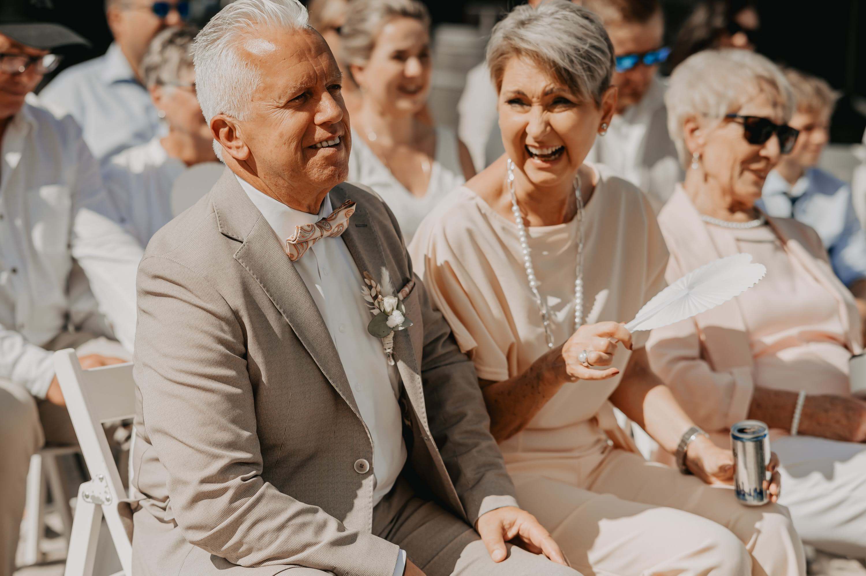 During the free wedding ceremony, the bride's parents sit next to each other, laughing and looking at each other.